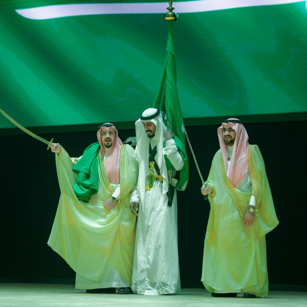 كل ثلاثة رجال تولى الغترة السعودية والعقال والشماع والبشت السعودي على التيراس، يلاحظ سيوفًا ويرفعون العلم السعودي احتفاءً باليوم الوطني. تتألق خلفية تهم بإضاءة خضراء مع ألوان العلم المرفرف فوقهم، في قوة قوية قصيدة عن اعتزاز جامعة القصيم بالوطن وه.
