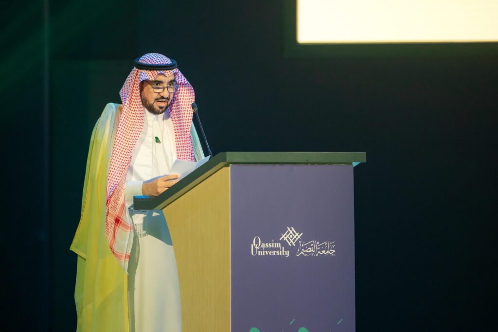 رجل الكت مرتدياً الغترة السعودية والعقال والبشت السعودي على منصة الخطابة التي تحمل شعار جامعة القصيم، ورئيساً أمام الحضور على خشبة المسرح، وذلك في حدود يطغى عليها إضاءة الخفتة وشاشة ساطعة في الخلفية، خلال فعاليات اليوم الوطني التي نظمتها جامعة القصيم.