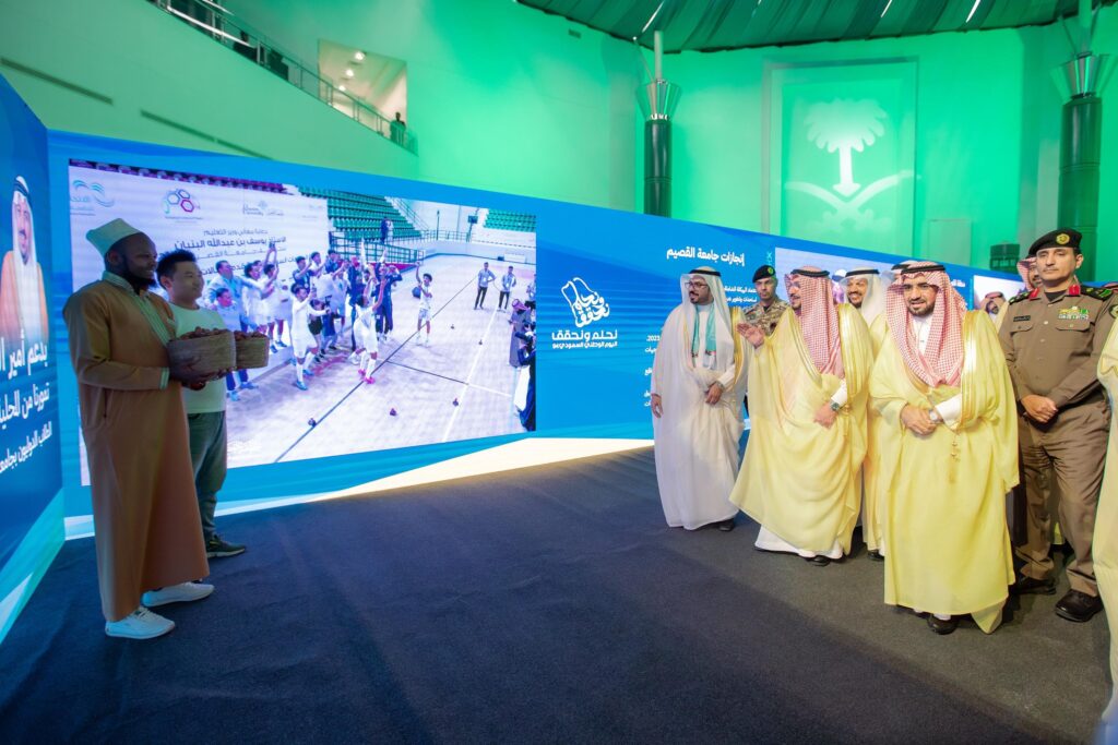 ويسير مجموعة من السعوديين مرتدين الغترة أو الشماغ السعودي، والعقال، والبشت السعودي، مرورًا بجانب شاشة رقمية كبيرة يعرض فعالية رياضية، وذلك داخل مقر مضاء بإضاءة ساطعة في جامعة القصيم. وتزدان القوس ببانرات معلوماتية زرقاء احتفاءً باليوم الوطني.