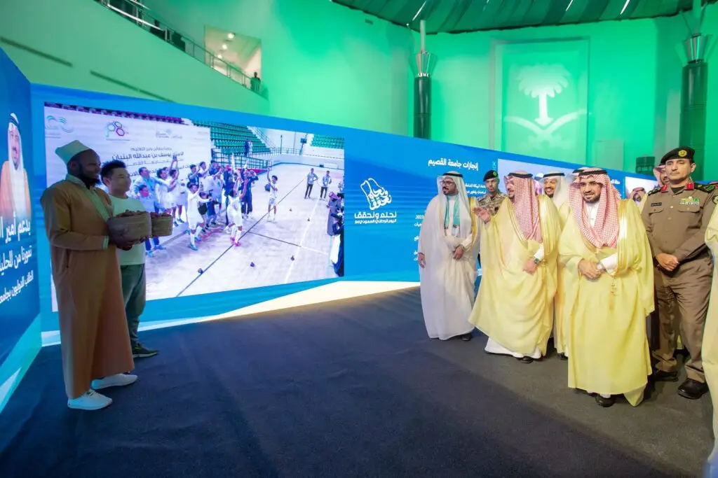 ويسير مجموعة من السعوديين مرتدين الغترة أو الشماغ السعودي، والعقال، والبشت السعودي، مرورًا بجانب شاشة رقمية كبيرة يعرض فعالية رياضية، وذلك داخل مقر مضاء بإضاءة ساطعة في جامعة القصيم. وتزدان القوس ببانرات معلوماتية زرقاء احتفاءً باليوم الوطني.