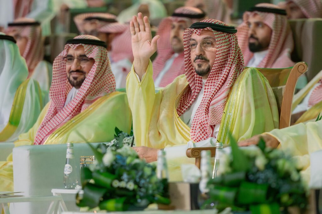 يجتمع عدد من الرجال ارتدين الغترة أو الشماغ السعودي مع العقال، في المنتصف رجل يرتدي البشت السعودي ييلوّح بيده اليمنى، بينما يتابع الحضور باهتمام. يبرز في مقدمة الصورة بعض المسطحات الخضراء وزجاجات المياه، في مشهد يعكس اليوم الوطني بمقر جامعة القصيم.
