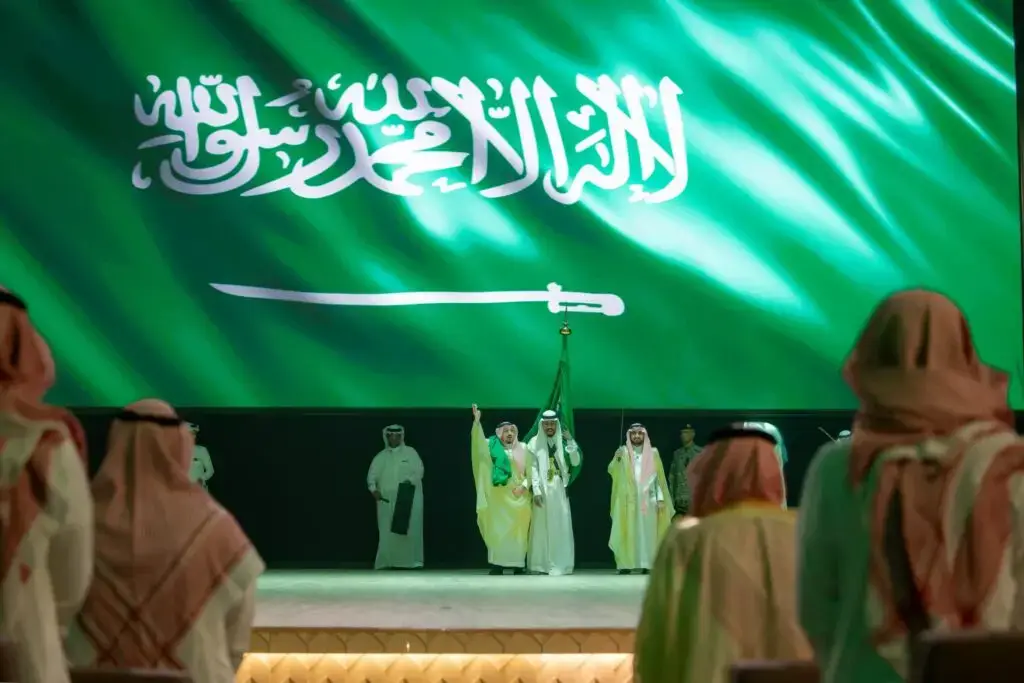 كات مجموعة من الأشخاص مرتدين الغترة أو الشماغ والعقال والبشت السعودي على منصة أمام شاشة كبيرة تحمل العلم السعودي، وذلك خلال ائتلاف اليوم الوطني الرابع والتسعين في جامعة القصيم، بحضور صاحب السمو أمير منطقة القصيم، بينما يتابع الحضور الحضور من الحضور.