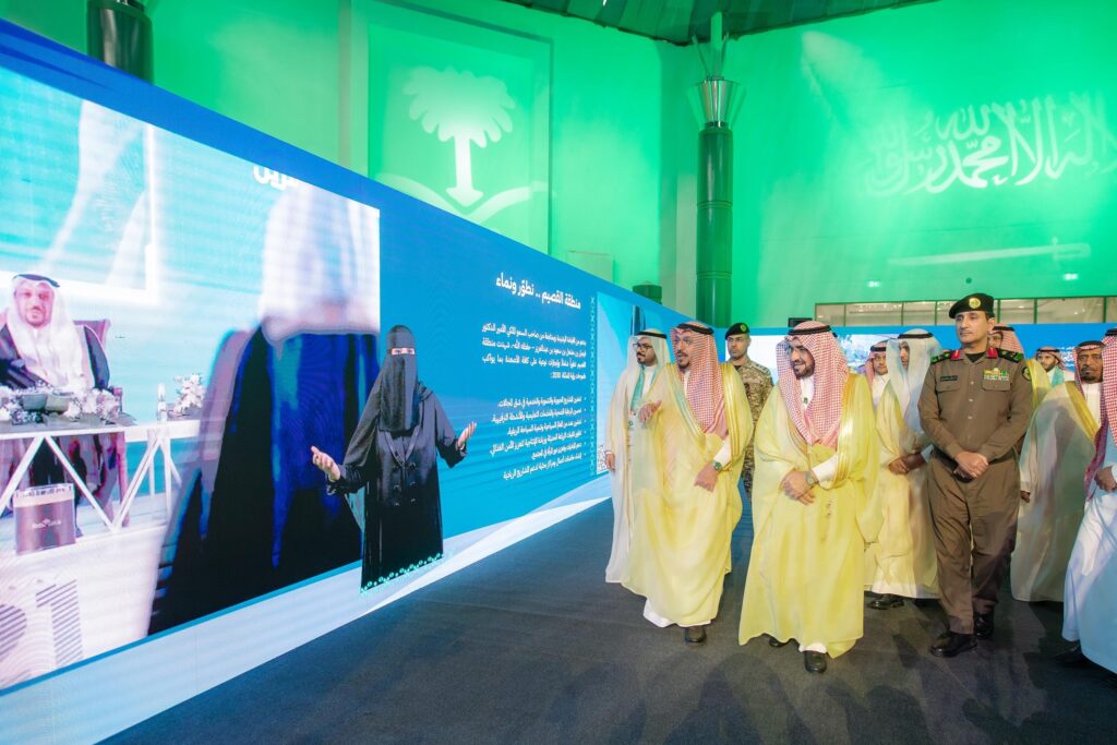 سار مجموعة من السعوديين، توقيع الغترة أو الشماغ والعقال والبشت السعودي التقليدي، طاقمة ضابط أمن، بجوار شاشة رقمية كبيرة رصدت سيدة فعالية، وتتزين الخلفية بإعلام المملكة العربية السعودية وإضاءة خضراء، وذلك بالتعاون بقوة باليوم الوطني في جامعة القصيم.