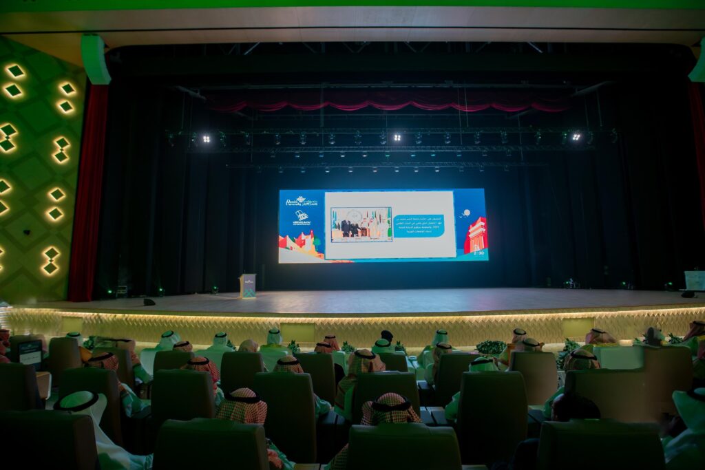 جلسة الحضور مرتدين الغترة أو الشماغ، والعقال، والبشت السعودي في قاعة الاحتفالات في القصيم، واجهون شاشة ضخمة تعرض عليها مادة العرض التقديمي. وقد أضفت إضاءة خضراء وعناصر عصرية للمناسبات المميزة احتفاءً بمناسبة اليوم الوطني.