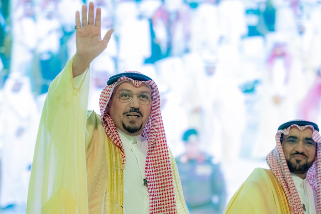يرفع أحد الرجال، مرتديًا الغترة السعودية والعقال والبشت السعودي المذهّب، يده ملوحًا خلال فعاليات اليوم الوطني في منطقة القصيم. ويقف بجانبه وعمه آخر يرتدي غترةقال وبشتاء فقط، بينما تتابع الحشود المحتفلة في الخلفية باهتمام.