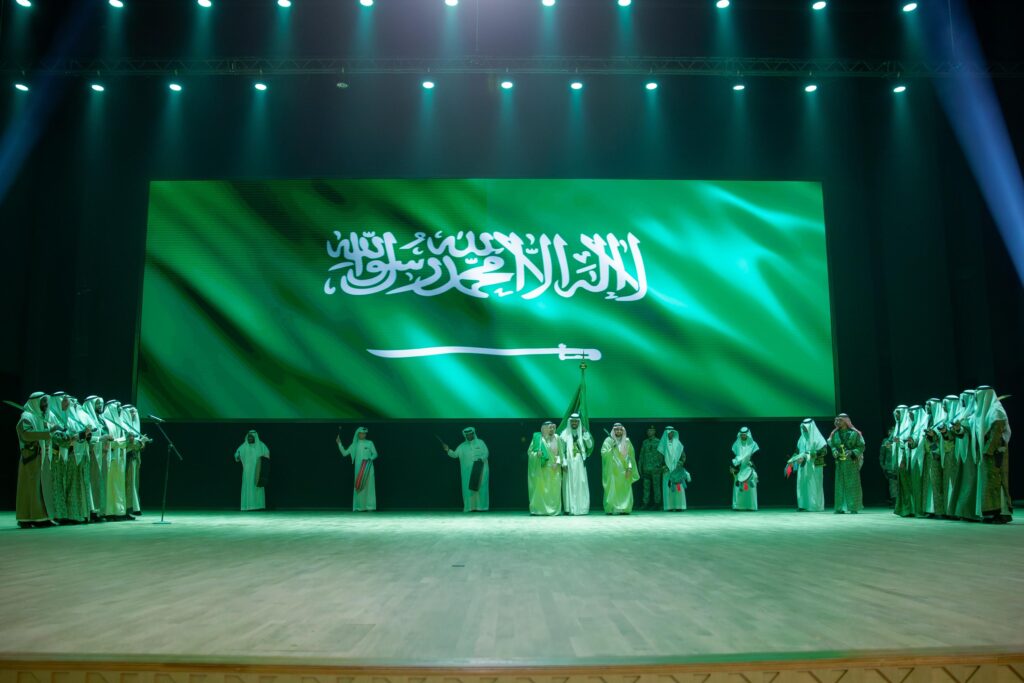 وقف جمع من الأشخاص مرتدين الغترة أو الشماغ والعقال والبشت السعودي على المسرح تحت شاشة كبيرة تزينها راية المملكة العربية السعودية، بينما أضاءت الأنوار الخضراء المكان خلال قوة اليوم الوطني في جامعة القصيم، وذلك بحضور صاحب السمو أمير منطقة القصيم.