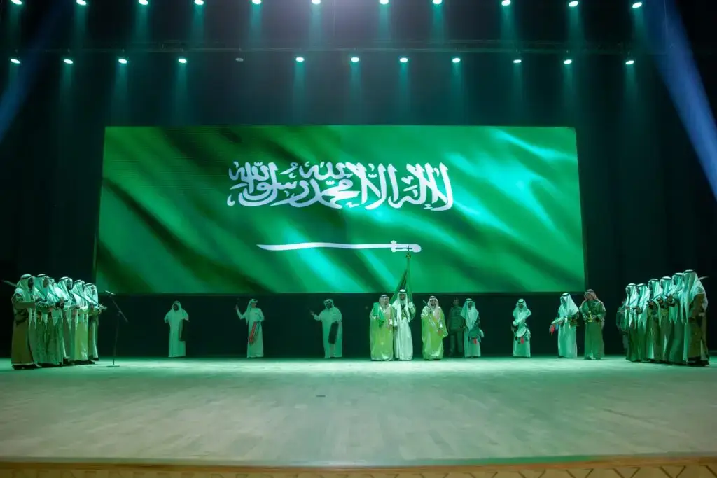 وقف جمع من الأشخاص مرتدين الغترة أو الشماغ والعقال والبشت السعودي على المسرح تحت شاشة كبيرة تزينها راية المملكة العربية السعودية، بينما أضاءت الأنوار الخضراء المكان خلال قوة اليوم الوطني في جامعة القصيم، وذلك بحضور صاحب السمو أمير منطقة القصيم.