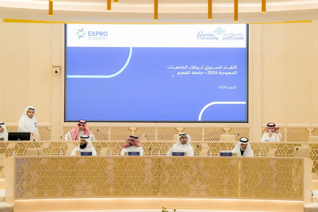يجلس ستة رجال غترة أو شماغاً وعقالاً وبشوتاً سعودية حول طاولة مؤتمر يعتمد بمابمايكروفونات، بينما اجتماع شخص مشهور على منصة الخطاب. في مراقبة شاشة كبيرة لبث نصًا عربيًا وشعاري "جامعة القصيم" و"وكلاء السعودية في الاجتماع".