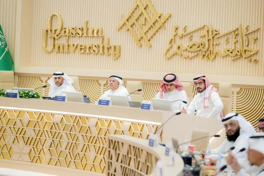 مجموعة من الرجال مرتدين الغترة أو الشماغ والعقال والبشت السعودي حول طاولة طعام طويلة تأكل بأجهزة حاسوب، وذلك أثناء حضورهم الاجتماع في جامعة القصيم. تسجيل خلفهم أعلام المملكة العربية السعودية واسم الجامعة باللغتين العربية والإنجليزية.