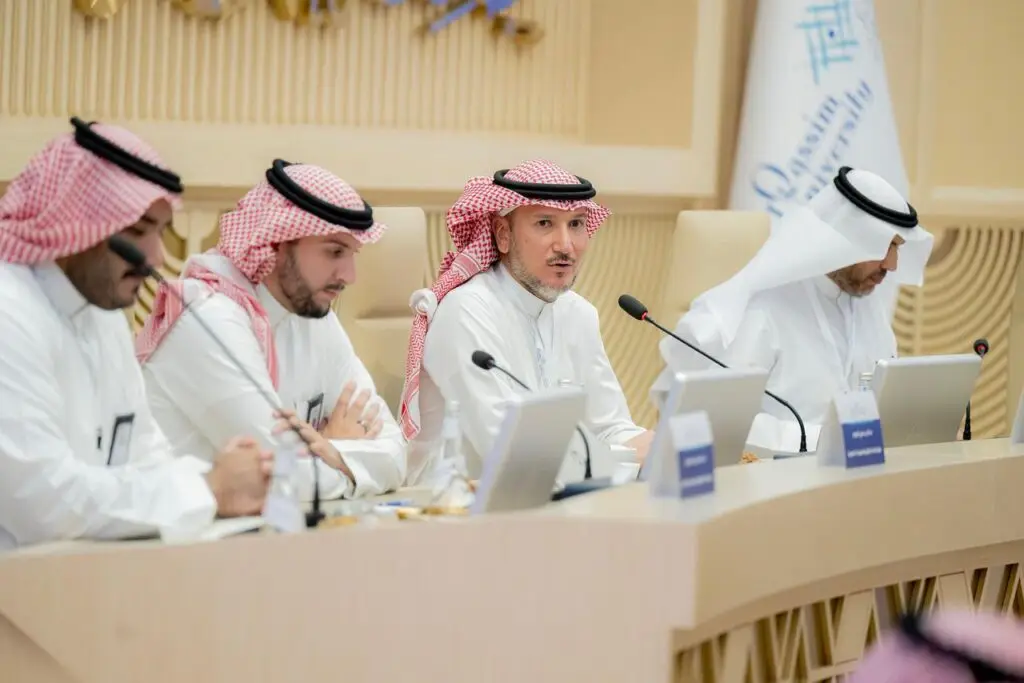 رجال يرتدون الغترة أو الشماغ السعودي مع العقال والبشتاء السعودي يجلسون حول أربعة طاولة مؤتمرات بالميكروفونات وأجهزة الكمبيوتر، ويشاركون في جلسة كمبيوتر نقاشية ضمن فعاليات الاجتماع المنزلي. ويظهر شعار جامعة القصيم في الخلفية، مما يبرز حضور وكلاء السعودية والأهمية الأكاديمية.