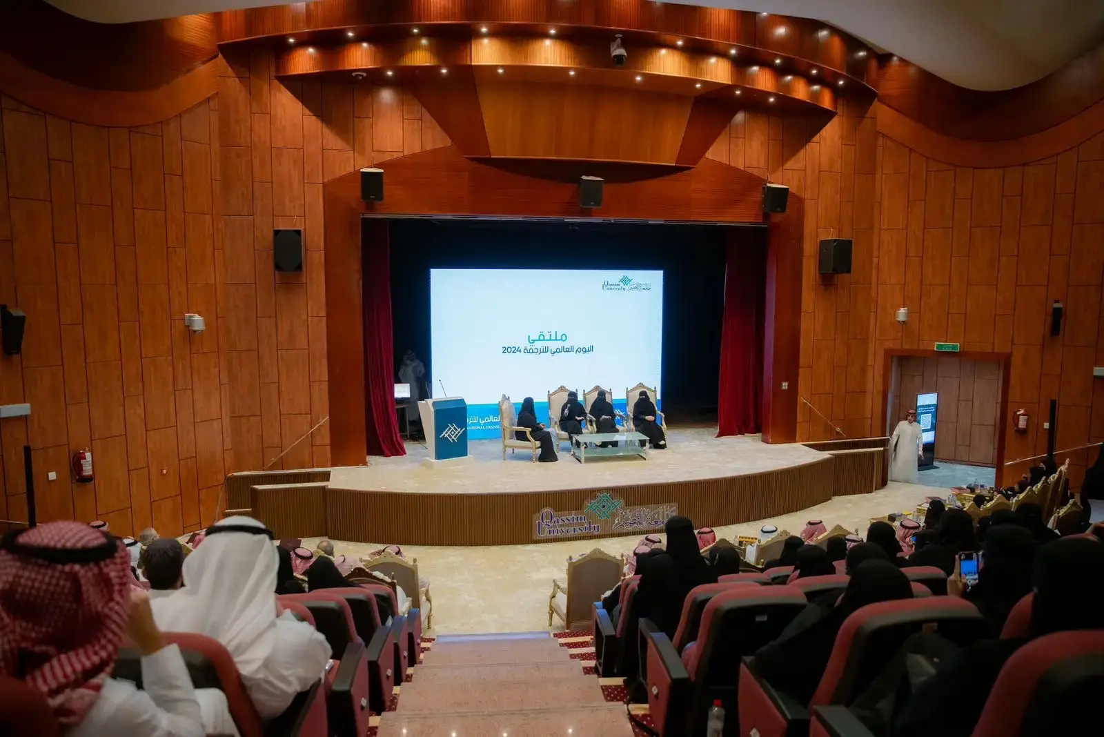 A special event to mark World Translation Day takes place in the specialized course with wood-paneled walls. A number of instructors from all languages sit on the podium, wearing the Saudi Ghutra or Shemagh and Egal with the Saudi Pasht, in front of a screen that automatically displays texts in Arabic, while the audience listens attentively and seriously to the theses presented at Qassim University.