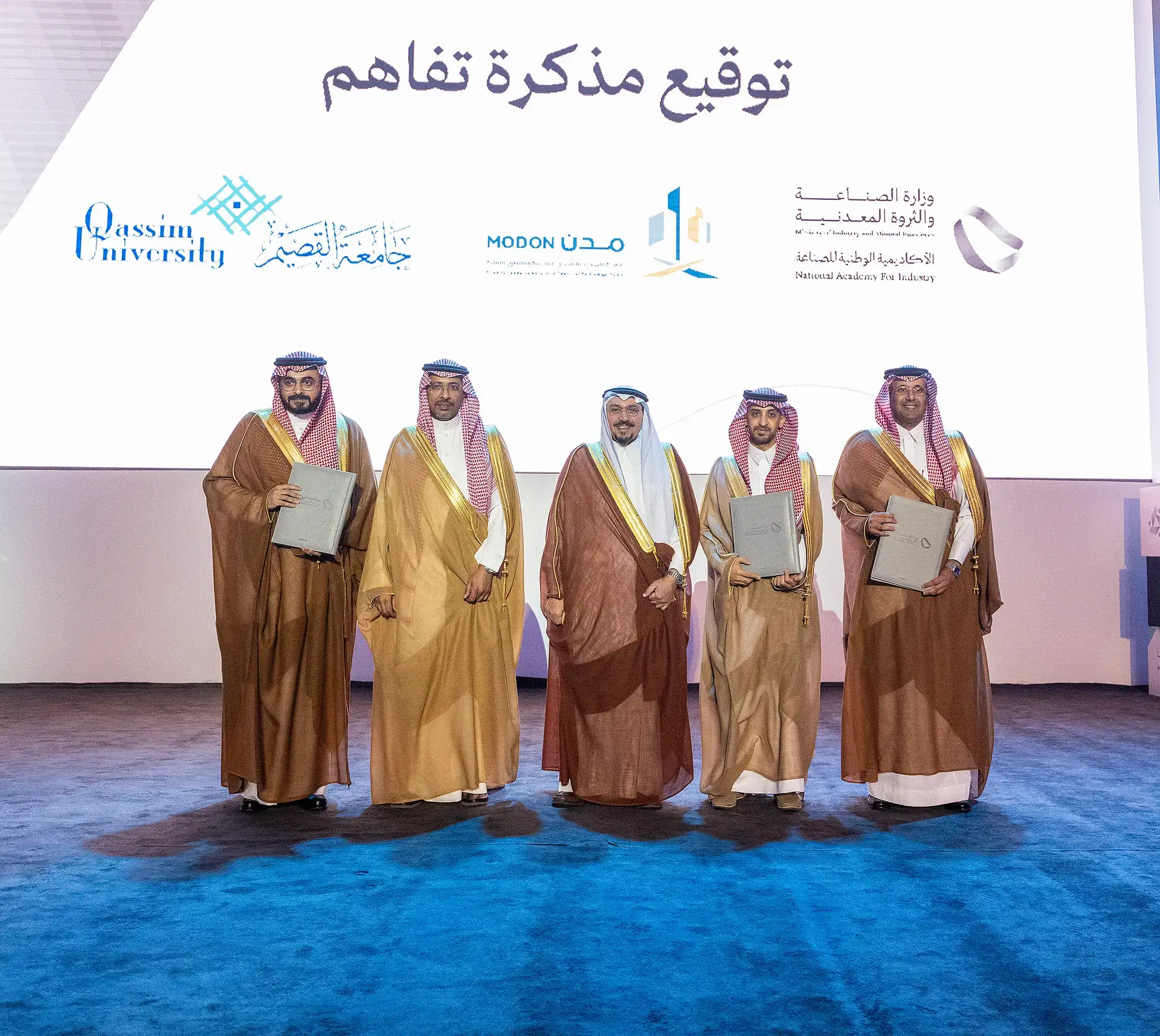 Five men wearing the Saudi ghutra or shemagh with an aqal, and some wearing the Saudi bisht, pose for a group photo in front of a background bearing the logos of Qassim University, the Saudi Industrial Cities Authority, the National Academy of Industry, and a number of other teams.