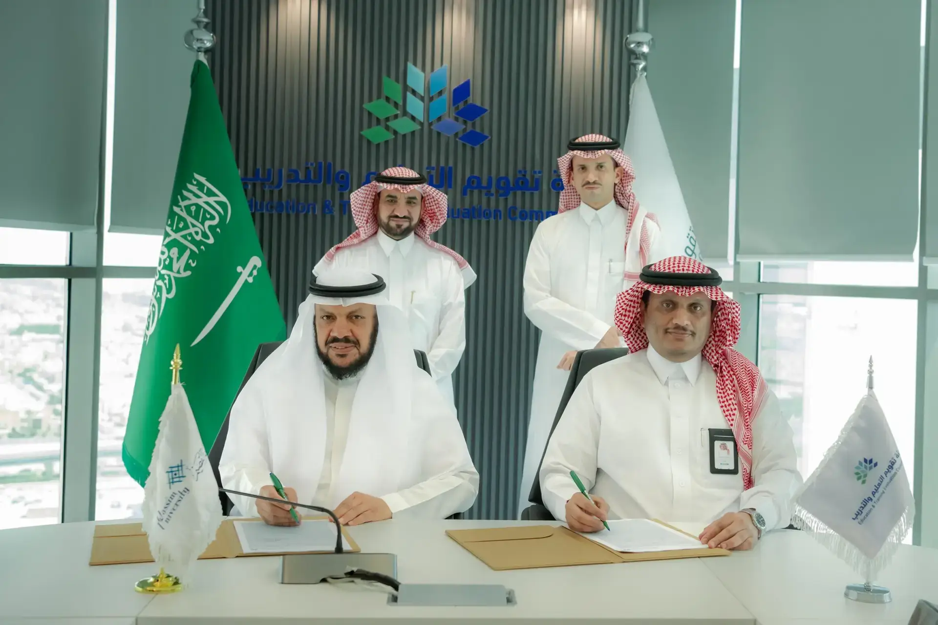 Men dressed in authentic Saudi dress in a Saudi besht, a Ghutrah or Shemagh, four in all, appear in the official office. Two of them speak and sign documents related to the accreditation of academic programs, while others communicate behind them. The flags of the Kingdom of Saudi Arabia and the Commission for Academic Excellence, as well as the university's logo and Arabic text, are depicted on the right side of the image.