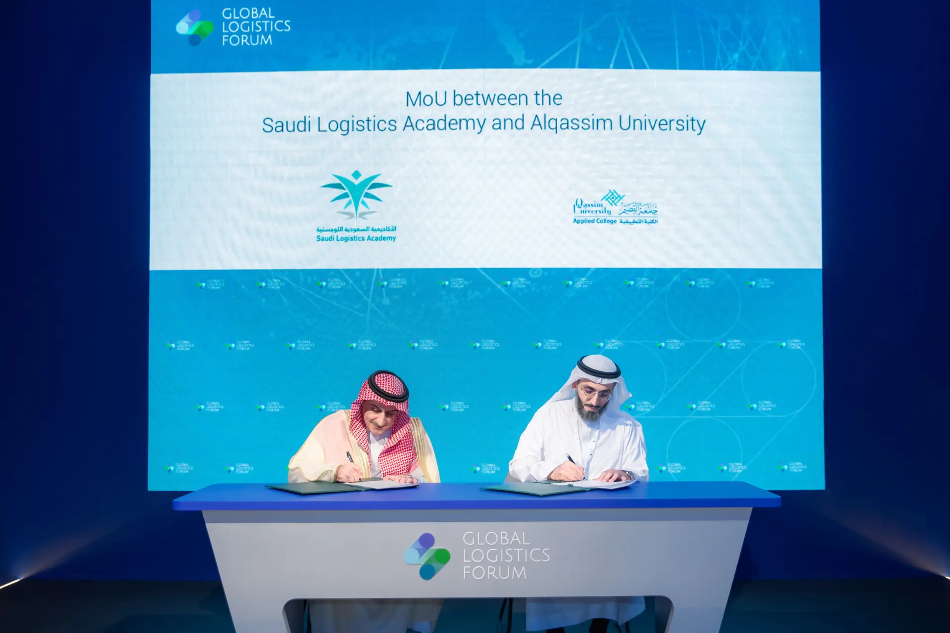 Two men dressed in Saudi Arabia's Ghutra, Eqal and Bisht signed a memorandum of cooperation through the Global Supply Chain Forum, with the screen behind them showing a partnership between the Saudi Academy and Qassim University.