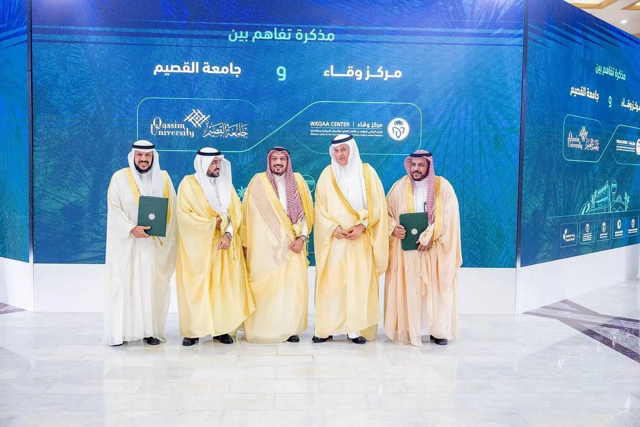 All five men in formal Saudi attire, wearing a shemagh or ghutra with an aqal and Saudi bisht, are gathered in an enclosed space, smiling and wearing plain women's clothing. Behind them is a blue background with texts and logos in Arabic for Qassim University and the national center "Waqa", on the occasion of the signing of a memorandum of understanding between the two organizations.