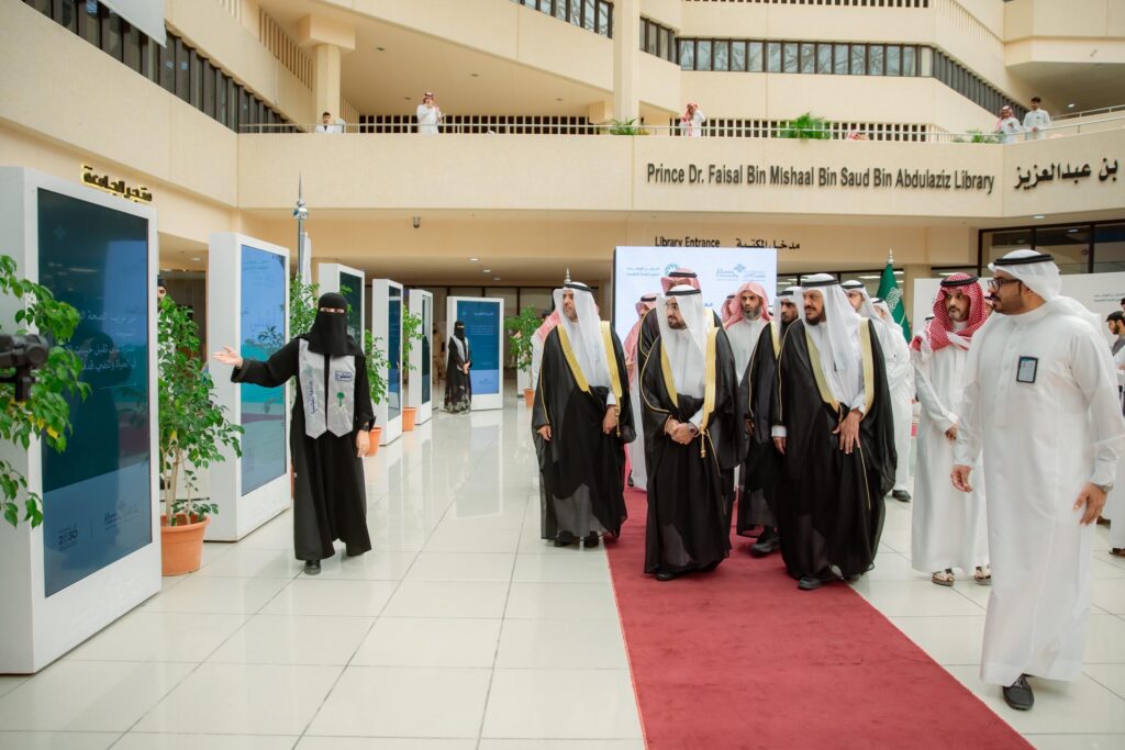 ويسير عدد من الرجال مرتدين الغترة أو الشماغ والعقال والبشت السعودي على السجادة الحمراء داخل أحد السجلات الحديثة، ومرفقة رئيس الجامعة وسيرتدي الحجاب ونقاباً أسود، وذلك خلال حضورهم معرض آمال لتعزيز الصحة النفسية. وتضفي الشاشات الرقمية والنباتات جمالاً وإشراقاً على الرجاء الفسيح.
