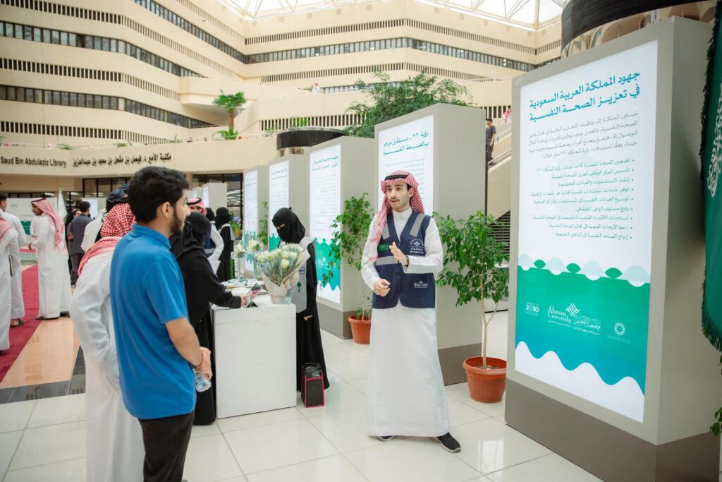 يتفاعل الحضور في معرض آمال مع نصوص رقمية واسعة النطاق تعرض نصوصًا باللغة العربية حول الصحة النفسية. ارتدى بعض المشاركين الشماغ السعودي أو الغترة السعودية مع العقال والبشت السعودي، بينما تواصل مع أحد أعضاء الفريق بجانب جناح يضم محتوى توعوية ومعلوماتية، وذلك في قاعة حديثة تابعة لجامعة القصيم.