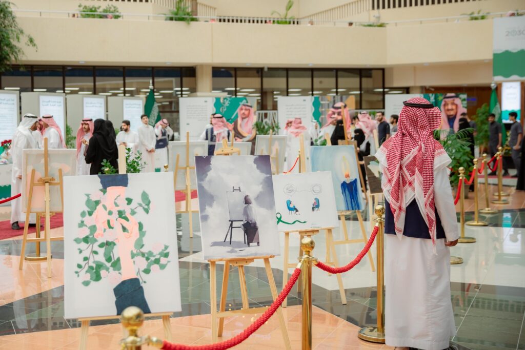 يجتمع مجموعة من الناس مرتدين الشماغ السعودي والسترة، مع مجموعة للقال والبشت الداخلي السعودي التقليدي، لمشاهدة معرض آمال الفنون في قاعة واسعة للمشاركة في القصيم. ولن يكون هناك إمكانية لإستخدام لوحات معروضة على حوامل ولوحات العلامات التجارية الحواجز الحمراء، وذلك في إطار مفاهيم الصحة الصحية الصحية.