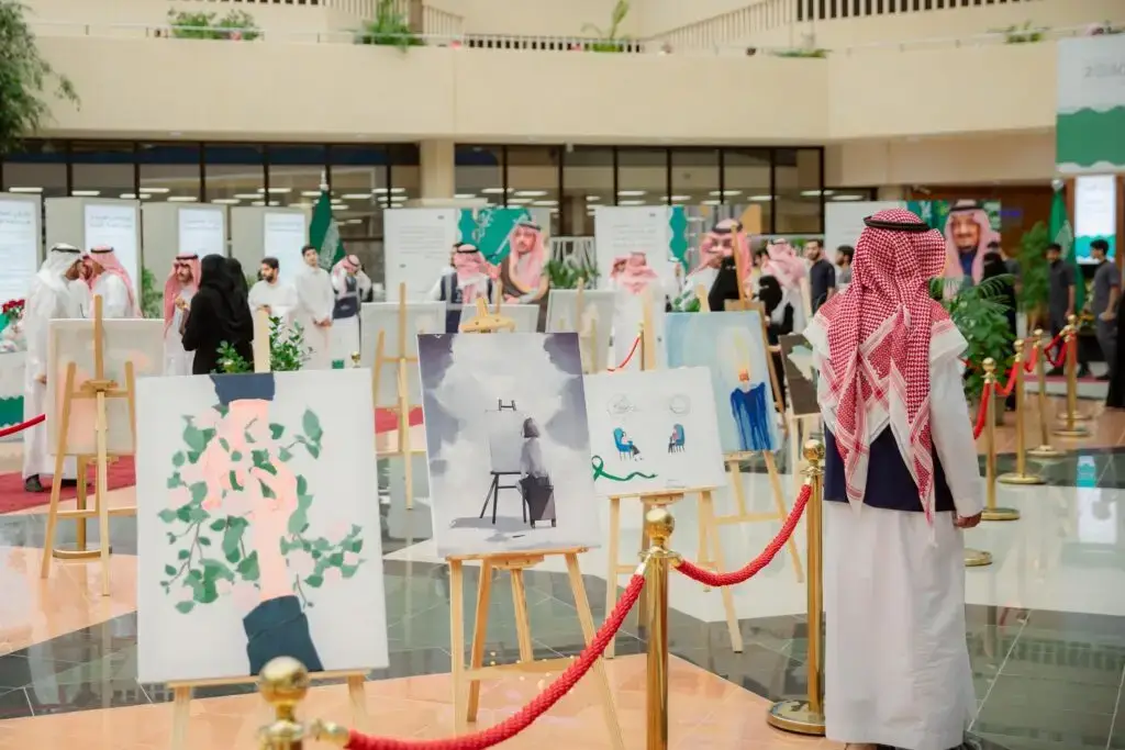 يجتمع مجموعة من الناس مرتدين الشماغ السعودي والسترة، مع مجموعة للقال والبشت الداخلي السعودي التقليدي، لمشاهدة معرض آمال الفنون في قاعة واسعة للمشاركة في القصيم. ولن يكون هناك إمكانية لإستخدام لوحات معروضة على حوامل ولوحات العلامات التجارية الحواجز الحمراء، وذلك في إطار مفاهيم الصحة الصحية الصحية.