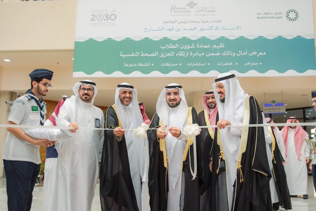 مجموعة من الرجال الحاضرين الغترة أو الشماغ والعقال والبشت السعودي، بالإضافة إلى الزي الرسمي، النشط بقص الشريط الأبيض خلال المباراة الرياضية في مكان مغلق. لأنها غير آمنة وبيضاء تحمل شعار "معرض آمال" لتعزيز الصحة النفسية باللغتين العربية والإنجليزية.