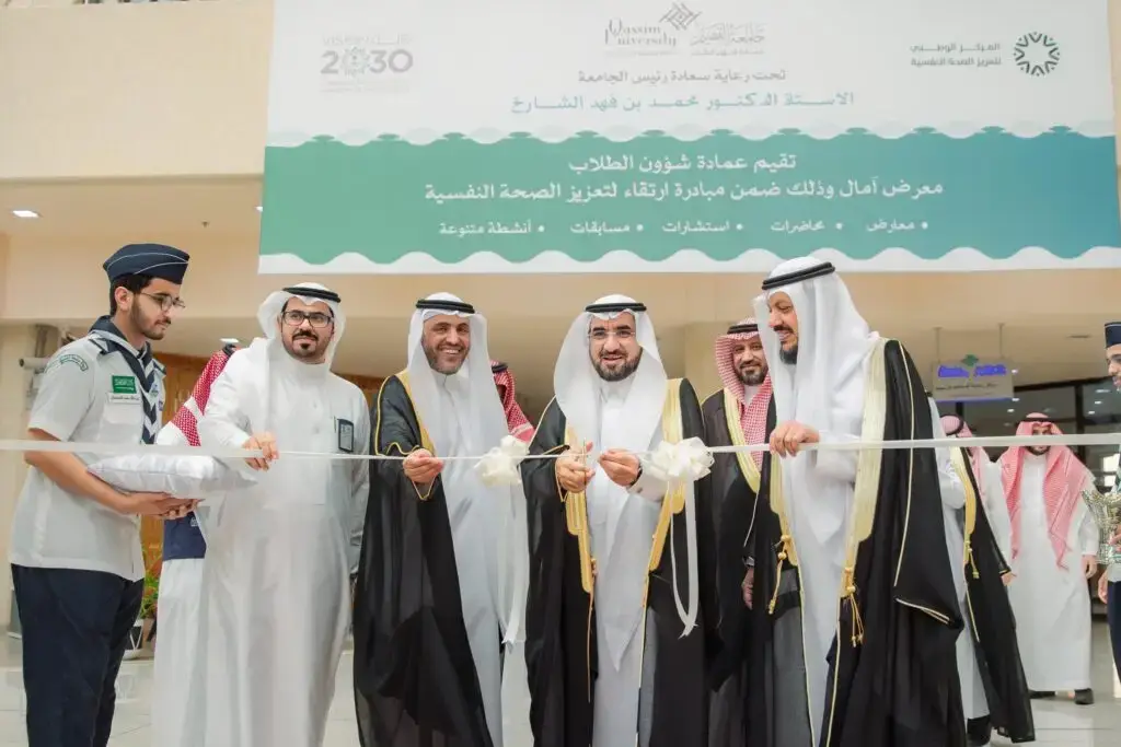 مجموعة من الرجال الحاضرين الغترة أو الشماغ والعقال والبشت السعودي، بالإضافة إلى الزي الرسمي، النشط بقص الشريط الأبيض خلال المباراة الرياضية في مكان مغلق. لأنها غير آمنة وبيضاء تحمل شعار "معرض آمال" لتعزيز الصحة النفسية باللغتين العربية والإنجليزية.