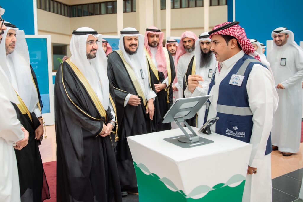 مجموعة من الرجال تولى الغترة السعودية والعقال والبشت السعودي في محيط كشك يستخدم رقمياً، داخل واحدة من فعاليات جامعة القصيم. ويحضر أمام الجامعة وهو يلقي كلمة حول أهمية الصحة النفسية، وذلك ضمن معرض آمال المقر في مقر الجامعة.