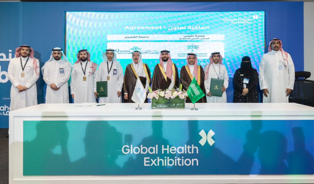 مجموعة الاتصال من الأشخاص، منهم الرجال الغتيرة أو الشماغ والبشت السعودي، إلى جانب امرأة، خلف طاولة تحمل أعلامًا ووثائق ضمن فعاليات معرض الصحة العالمي. يبرز في الخلفية شاشة تجريبية كبيرة باللون الأزرق، في نموذج تأكيد التعاون العلمي بين الحضور ويركز على تبادل الخبرات والدراسات الطبية بين جامعة القصيم والمؤسسات المشاركة.