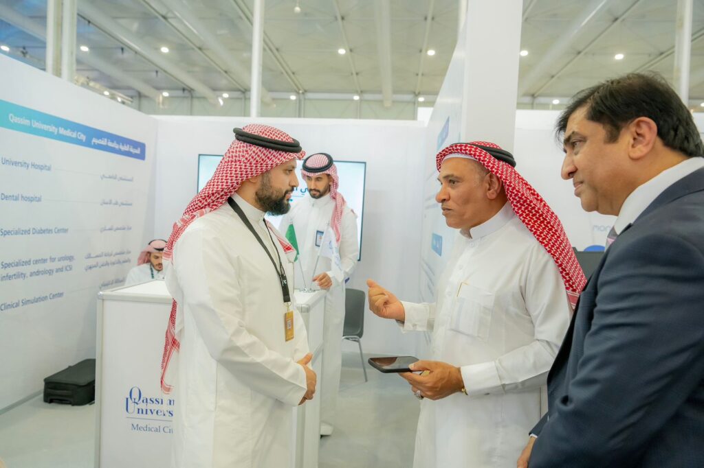يتبادل مجموعة من الرجال الذين ينتظرون البشتاء السعودي والغترة والعقال، بالإضافة إلى قمصان الراعي النازية، الخبرات حول الدراسات الطبية داخل جناح المعرض. عصر أحدهم جهازاً لوحياً بينما يغغي الآخر باهتمام وسط الشاشات التعريفية واللوحات المعلوماتية الخاصة بجناح جامعة القصيم.