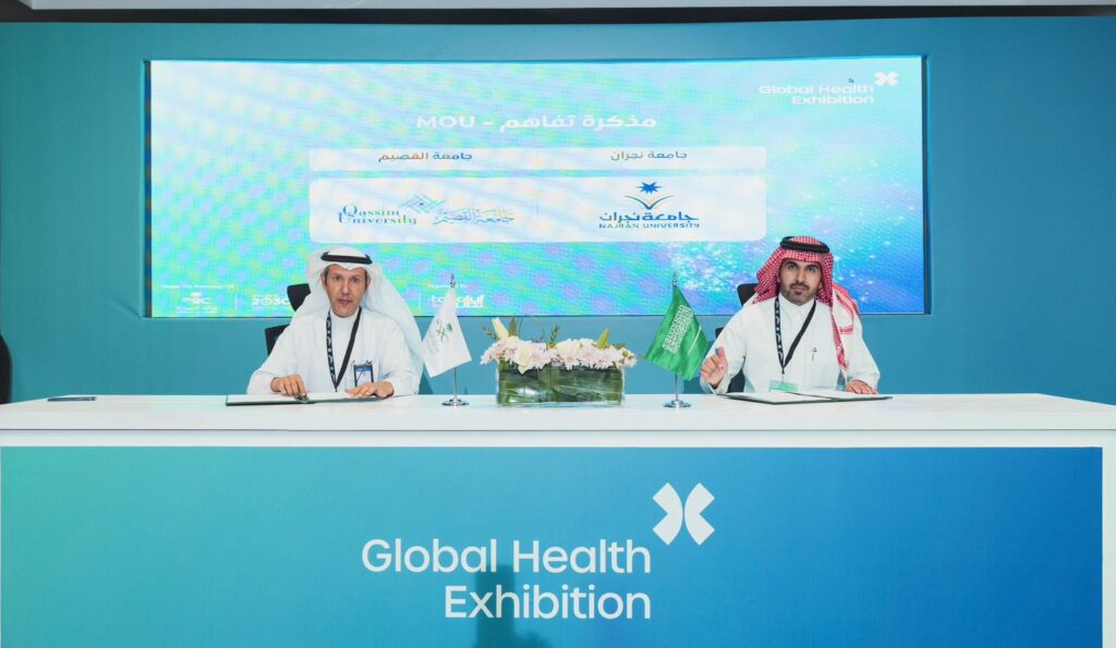 يجلس رجلان يرتديان الغترة والعقال والبشتاء السعودي حول طاولة بميكروبات وأعلام، وينظمان توقيع مستندات خلال فعاليات المعرضة الصحية العالمية، وذلك بالتأكيد على تحسين تبادل الخبرات والدراسات والبحوث الطبية. اعتماد اعتمادات المطبوعات والنصوص الرسمية على الشاشة خلفهما.