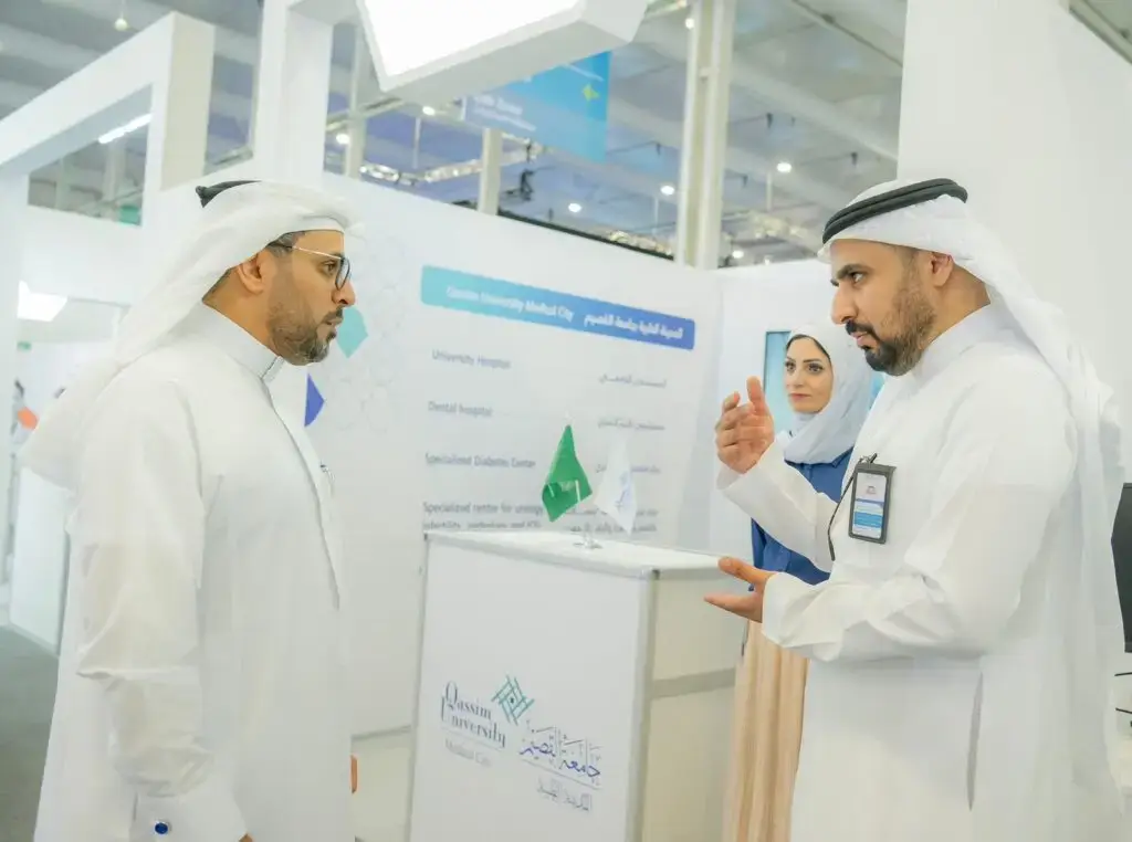 Three famous old Saudi Arabian men, from 15 Ghutrah, Shamakh, Aqal and Bashta, chatting at the event booth of the Medical Academy at Qassim University. One of the men is shown explaining with his hand during the conversation, while the other man listens intently to the woman, standing nearby. The pavilion's platform was utilized for medical information in both Arabic and English.