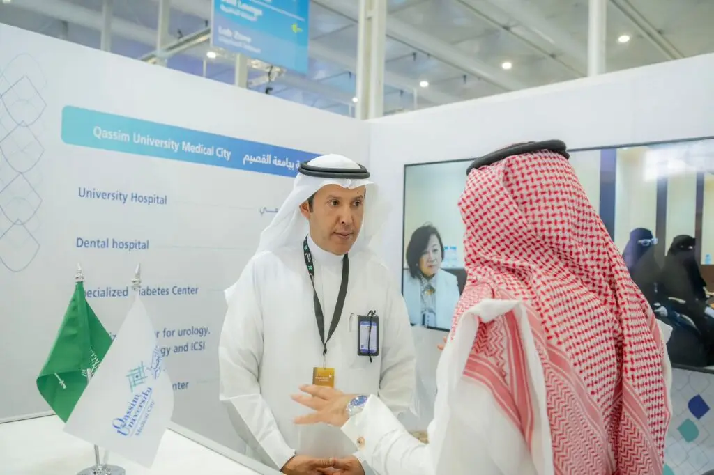 Two men dressed in traditional Saudi attire (thobe with shemagh or Saudi ghutra) chat at the Qassim Medical City pavilion, where Saudi Arabia's flags and informational signs were on the counter. One of the women's screens highlighted the importance of exchanging experiences in medical studies and research.
