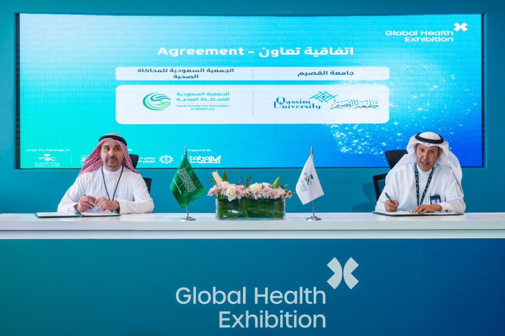 يجلس الرجلان ويرتدي الغترة والشماع والعقال والبشت السعودي على طاولة للتوقيع بشكل تعاوني ضمن فعاليات المعرضة الصحية العالمية، ويركز التعاون على تبادل الخبرات والدراسات والبحوث الطبية. توصية في شاشة تحمل شعاري جامعة القصيم والجمعية السعودية للملاحظة في الرعاية الصحية.