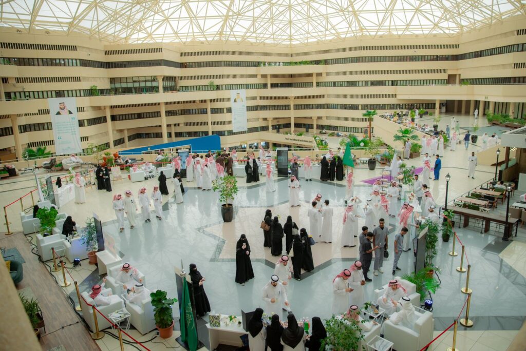 قاعة داخلية نسائية واسعة وسقوف عالية في جامعة القصيم، تحتضن رجال مرتدين البشوت السعودية والستر أو الشماغ والعقال، والسيدات بزيهن التقليدية. يتجمعون حاضرين حول الطاولات والأركان الموزعة في الأتريوم الهندسي الحديث، حيث تمثل الهيئات الحكومية وتبحث عن فرص الشراكات المجتمعية بما في ذلك الأصالة والهوية الوطنية.