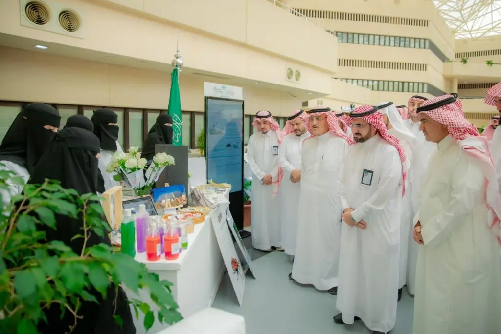 اجتمعت مجموعة من الرجال مرتدين الثياب البيضاء مع الشماغ أو الغترة السعودية، مرتديات العبادات والنقاب الأسود، حيث طاولة عرض تحتوي على مواد تعريفية، وذلك في إطار إبراز الشراكات المجتمعية وبحضور الممتازة رئيس جامعة القصيم. تأليف المملكة العربية السعودية في القاعة ذات الإضاءة الساطعة.