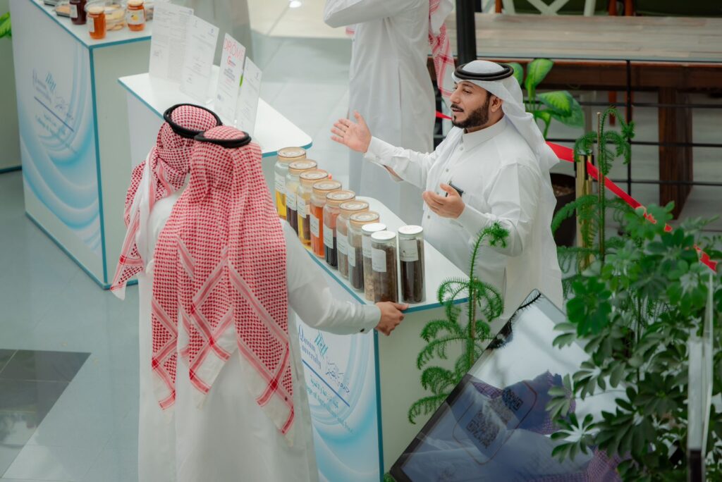 يتبادلان رجلان يرتديان الغترة والعقال والبشتاء السعودي الحديث عند منصة تعرض عدداً من أوعية عشبية أو عشبية، في مشهد يسلط الضوء على الشراكات المجتمعية. وتوجد لوحات تعريفية بالمعلومات خلف المنصة، كما يظهر في مقدمة الصورة الشخصية ما لا يقل عن عدد من النباتات.