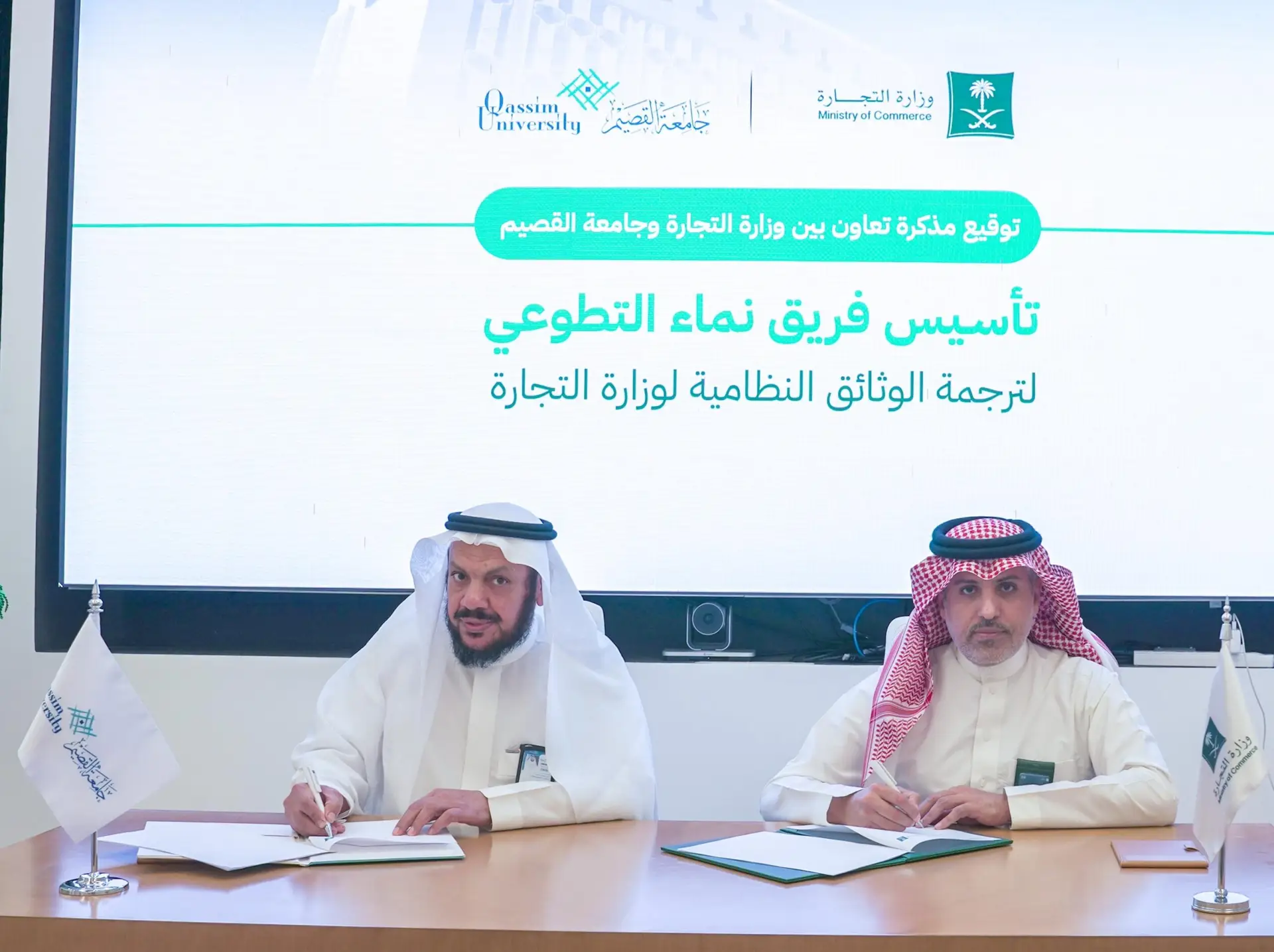 Two men wearing the Saudi shemagh sit at the duo's table and sign the memorandum of cooperation. Behind them is a text screen in Arabic and the logos of Qassim University and the Ministry of Commerce. Small flags are also placed on the table.