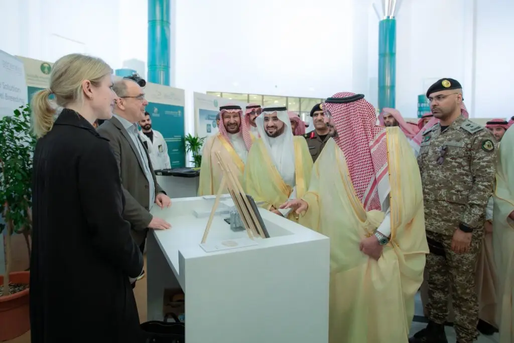 تواجد مجموعة من الرجال الحاضرين الزي السعودي التقليدي في الشماغ أو الغترة، إلى جانب أحد الجنود، حيث كانوا يتحاورون مع شخصين في منصة داخل قاعة المعرض، وذلك خلال الفعاليات العلمية الدولية الثانية للإيبل. في خلفية الشاشة عدد من الملصقات التعريفية والنباتات، مما يعكس الطابع التنظيمي والمحتوى العلمي الذي حصل على النتيجة تحت إشراف جامعة القصيم.
