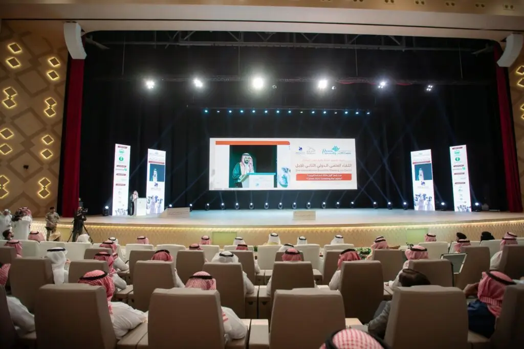 قاعة المؤتمرات بحضور حضور الزي السعودي التقليدي، ويواجهون منصة مضاءة بشكل ساطع خلال فعاليات "اللقاء العلمي الدولي للإبل" الذي تنظمه جامعة القصيم؛ حيث تتزين المنصة بلافتات خاصة، ويتحدث أحد أفرادها مرتدياً حفلة وشماغاً أو اغترة سعودية.