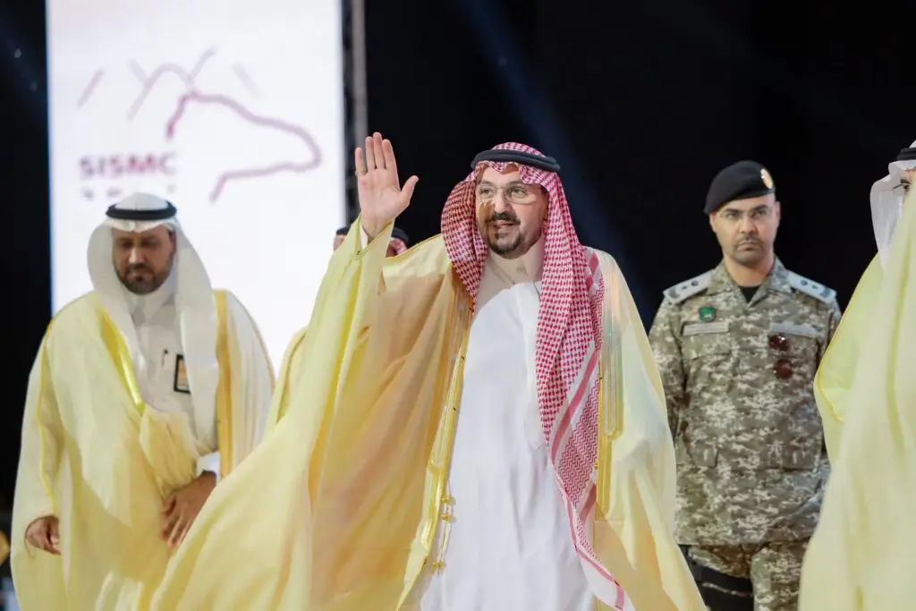 يظهر في الصورة رجل يرتدي الزي السعودي التقليدي، يشمل الشماغ أو الغترة السعودية، يلو بيده أثناء سيره وبجواره عدد من الأشخاص، من بينهم أحد أفراد القوات العسكرية. وفي الخلفية شاشة كبيرة تعرض عليها كتب باللغة العربية وشعار "اللقاء العلمي الدولي للإبل" في جامعة القصيم.