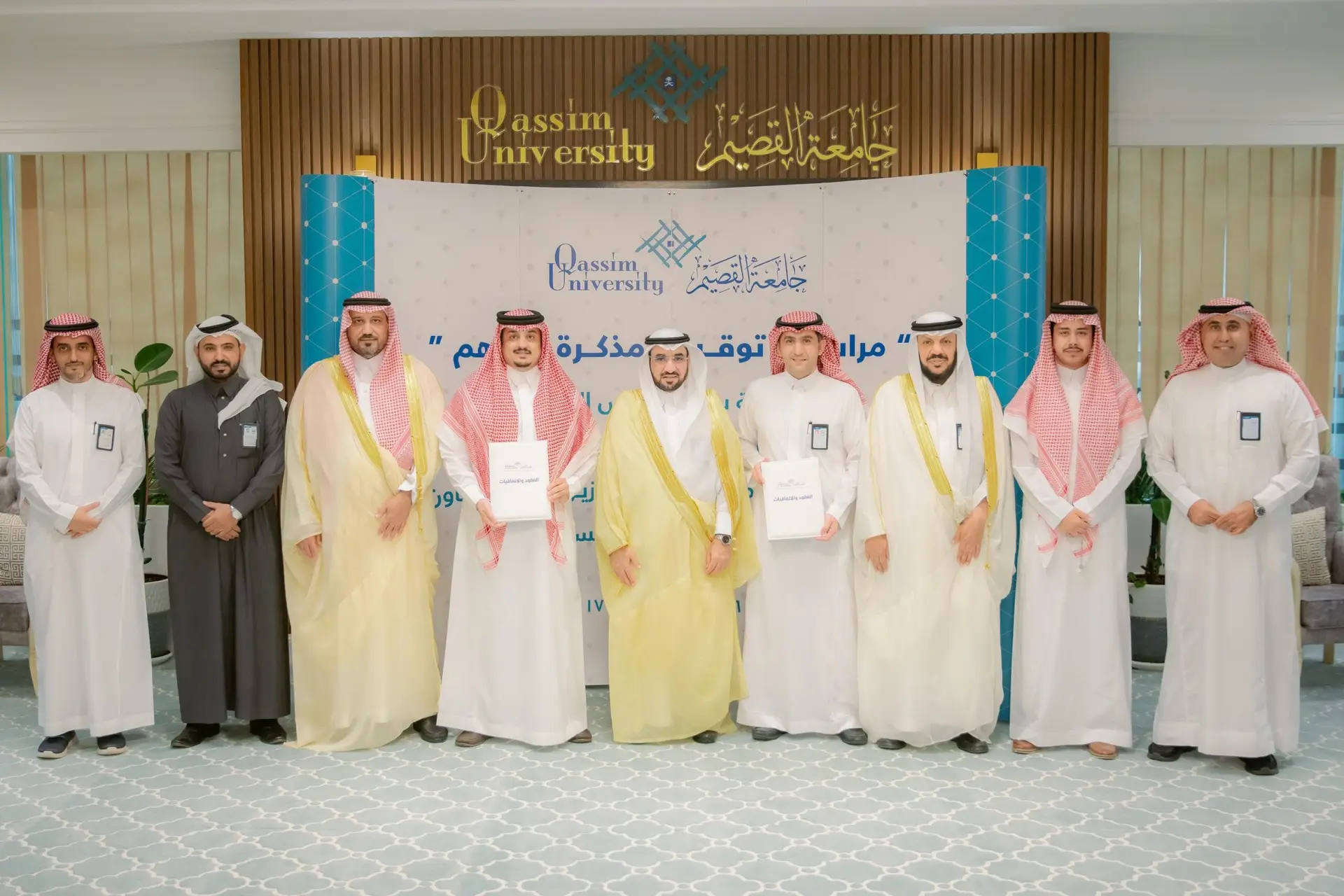 A number of men wearing Saudi shemaghs, Saudi ghutrahs, uniforms, and suits are lined up in a closed space, and a group photo in front of you bears the logo of Qassim University and the sign of the College of Business and Economics. You appear in the center of the photo twice.