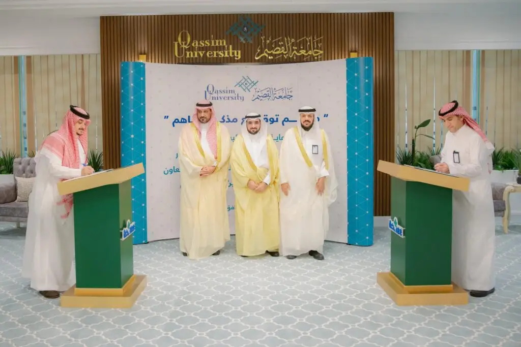 Five men in authentic Saudi attire, including a shemagh or Saudi ghutra, call into the hall as two of them sign a memorandum at the podiums. Noticeable behind them is the logo of Qassim University, highlighting a partnership between an all-ages, all-professional business school endorsement company.