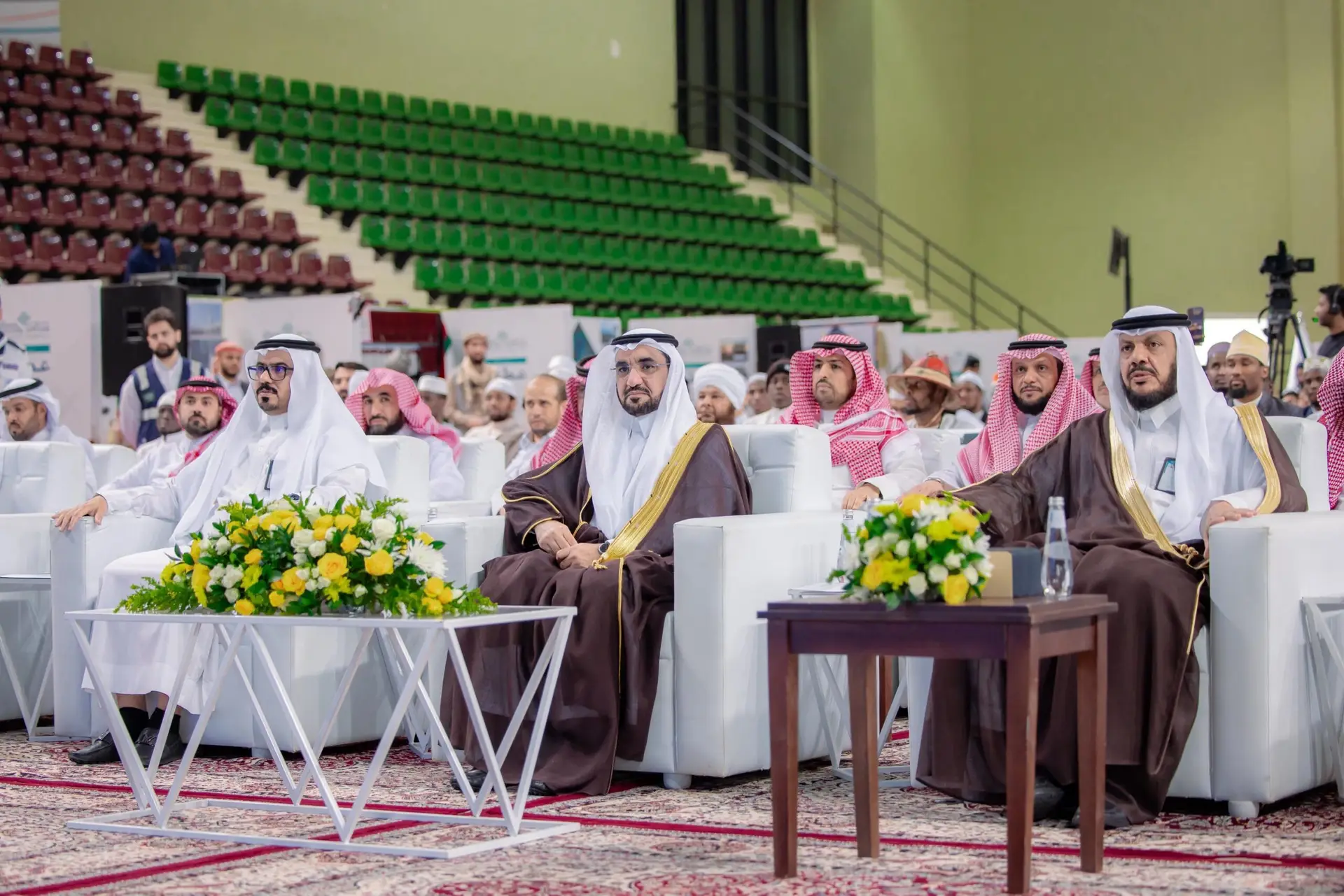 يجتمع عدد من رجال مرتدين الزي السعودي الرسمي، بما في ذلك الشماغ أو الغترة السعودية، على كراسٍ ثلاثة خلال فعالية عطاء في القصيم. وتزين الجداول تنسيقات الزهور، بينما تعيش في الخلفية المدرجة بقاعدة خضراء وحمراء.