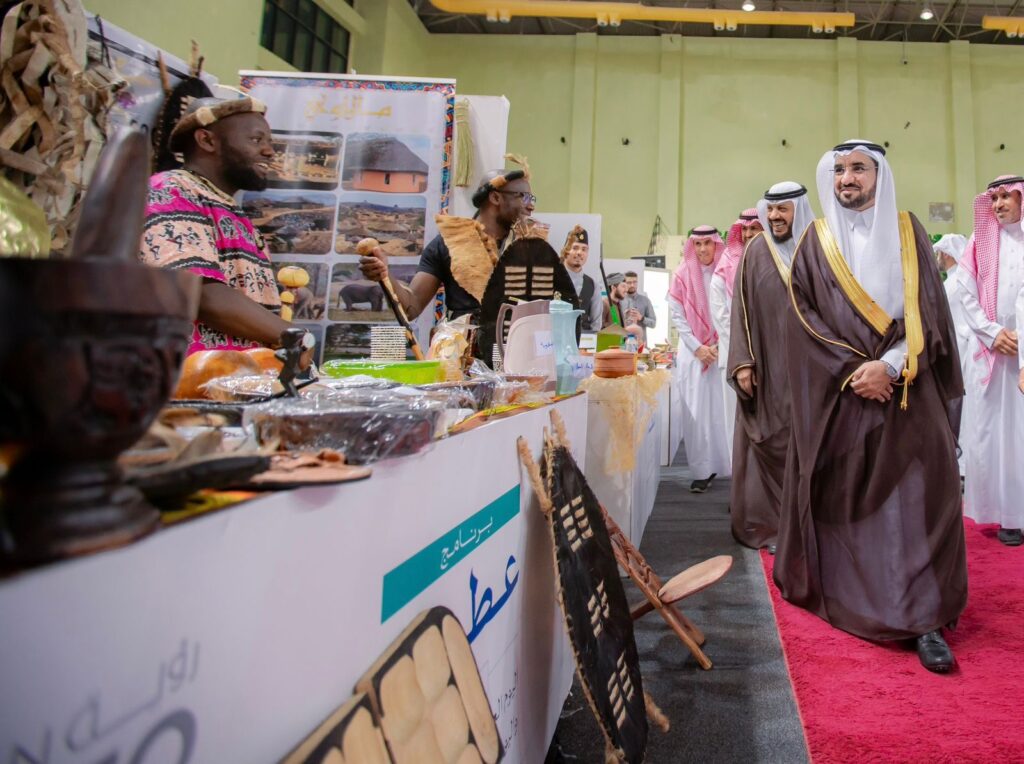 يمر مجموعة من الرجال تولى الغترة أو الشماغ والعقال والبشت السعودي بعد جناح معرض يُعرض فيه منتوجات ومأكولات أفريقية ثقافية، بينما تتصل للطلبات الدوليةون، المشاركين في برنامج عطاء، خلف مرتدين أزياء أفريقية زاهية الألوان.