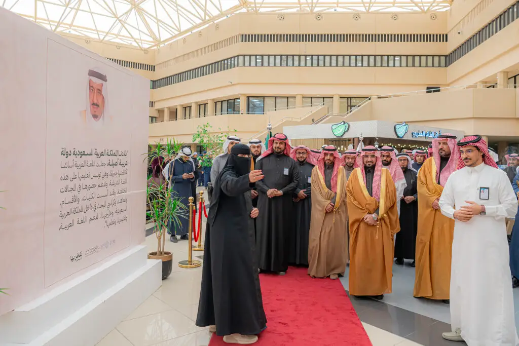 تجمّع عدد من الرجال بزيهم السعودي الأصلي، الحضور الشماغ أو الغترة السعودية، داخل قاعة عالمية بالقرب من سجادة حمراء في إطار نظمها الفعالة جامعة القصيم احتفاء باليوم للغة العربية. واستمع إلى كلمة ألقاها أحد الوجهين، بينما يأتي على الجانب المعماري لوحة عرض كبيرة تتضمن نصوصًا باللغة العربية وصورة شخصية، وذلك من خلال وسط المدير المعماري.