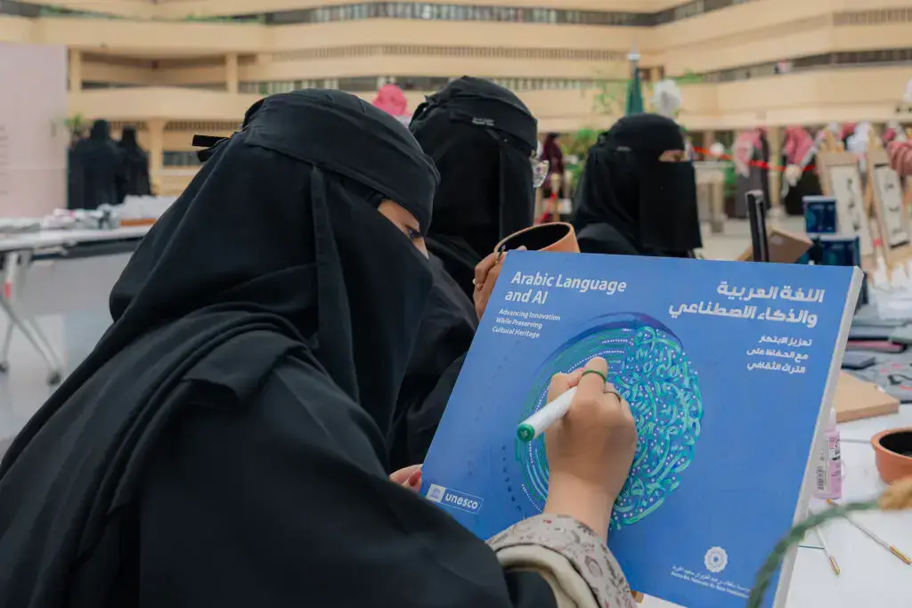 ترتدي نقاباً وشماغاً أو غترة سعودية في طيران الخليج تحت عنوان "العربية والذكاء الاصطناعي"، وذلك على طاولة أربعة أخريات ترتدي زياً مماثلاً، خلال الاحتفال باليوم العالمي للغة العربية، وذلك في قاعة متعددة الحضور في القصيم.