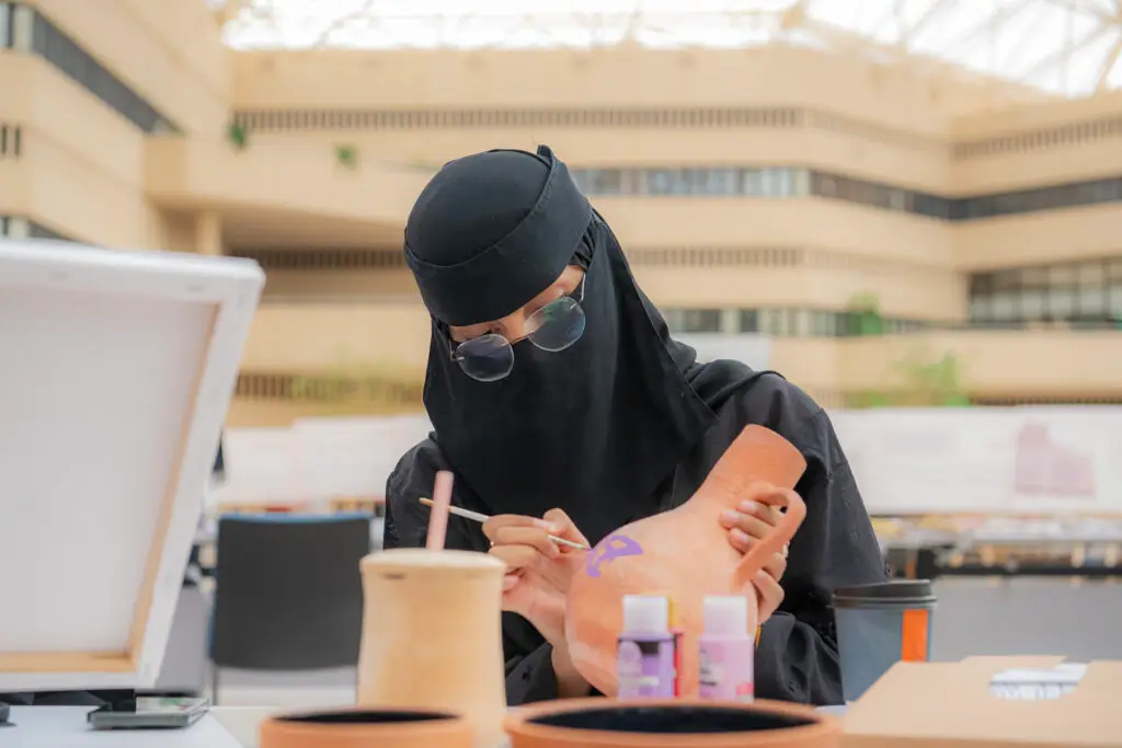خلال فعاليات الفعالية وتحديداً اليوم العالمي للغة العربية في جامعة القصيم، بذلة نسائية ترتدي ملابس سوداء ونقاباً ونظارة شمسية بتلوين مزهرية فخارية البنفسجي في قاعة الملابس الداخلية الرائعة. وقد أحاطت به تقنية قطع من الفخار المتنوع، مما يضيف إلى المكان طابعاً إبداعياً وثقافياً وحقيقياً وروحياً.