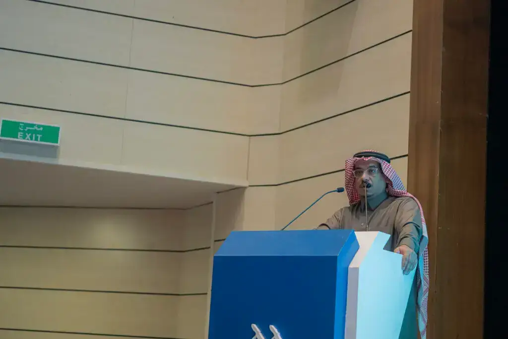 رجل يتحدث مرتدياً الثوب والشماغ السعودي في منصة الخطابة في قاعة الحديث في القصيم، وخلفه أخضر لمخرج الطوارئ، وذلك عبر فعالية اليوم العالمي للغة العربية.