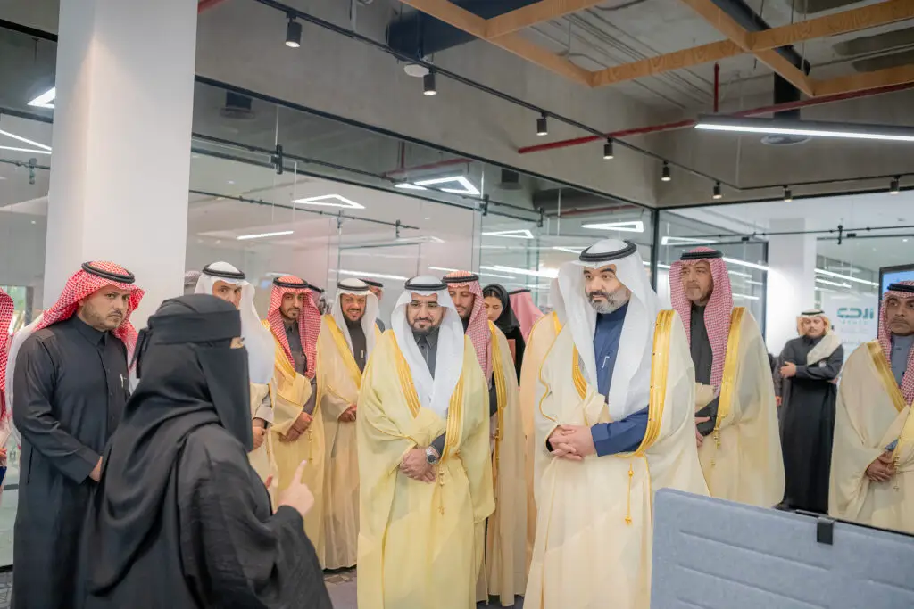 يجتمع مجموعة من الرجال مرتدين الثياب الرسمية مع الشماغ السعودي أو الغترة السعودية، يستمعون باهتمام إلى سيدة ترتدي ملابس سوداء ونقاباً، وذلك داخل مكتب حديث ذي بلاط في جامعة القصيم، حيث تتميز بإضاءة ساطعة وتصاميم فنية في هندسية.