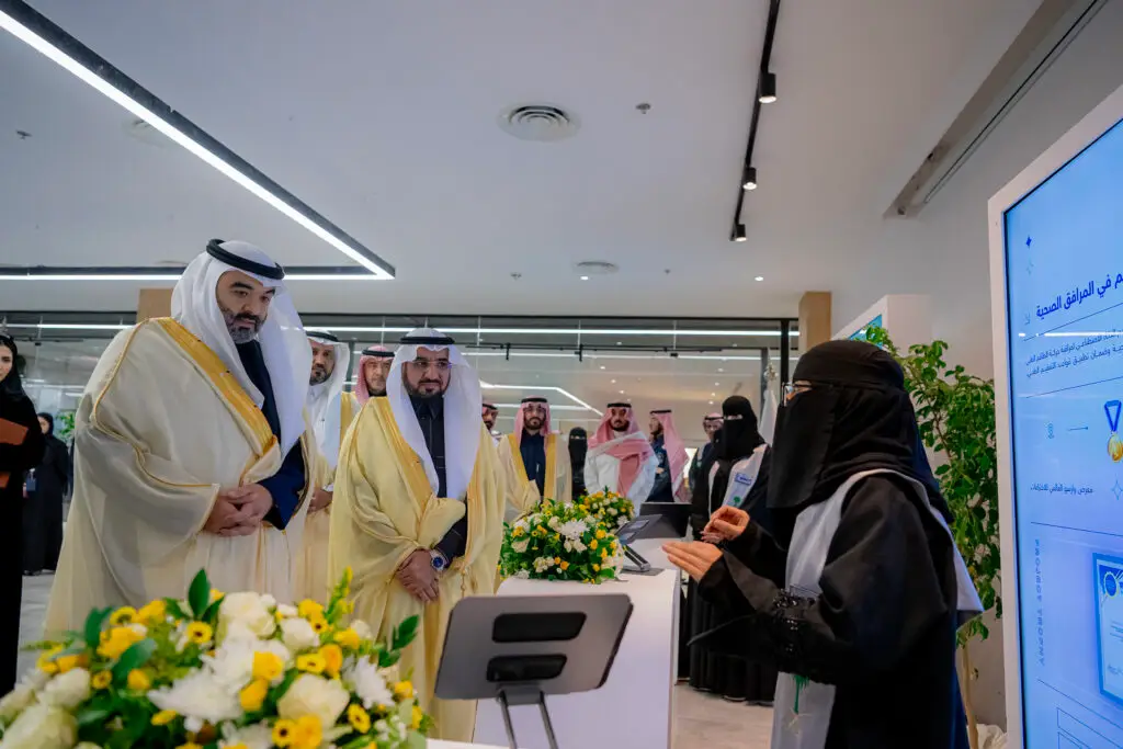 يجتمع مجموعة من الرجال مرتدين الشماغ أو الغترة السعودية مع الثوب التقليدي، وهم يستمعون باهتمام إلى سيدة تقدم عرضًا في إحدى المنصات التقنية وهي ترتدي أداءة ونقاباً الأسود، حيث تقوم باستعراضها للبحث المباشر. ضع في اعتبارك المكان الحديث من الزهور وشاشة رقمية كبيرة، ما يعكس الطابعة المتطور للفعالية المقامة في جامعة القصيم.