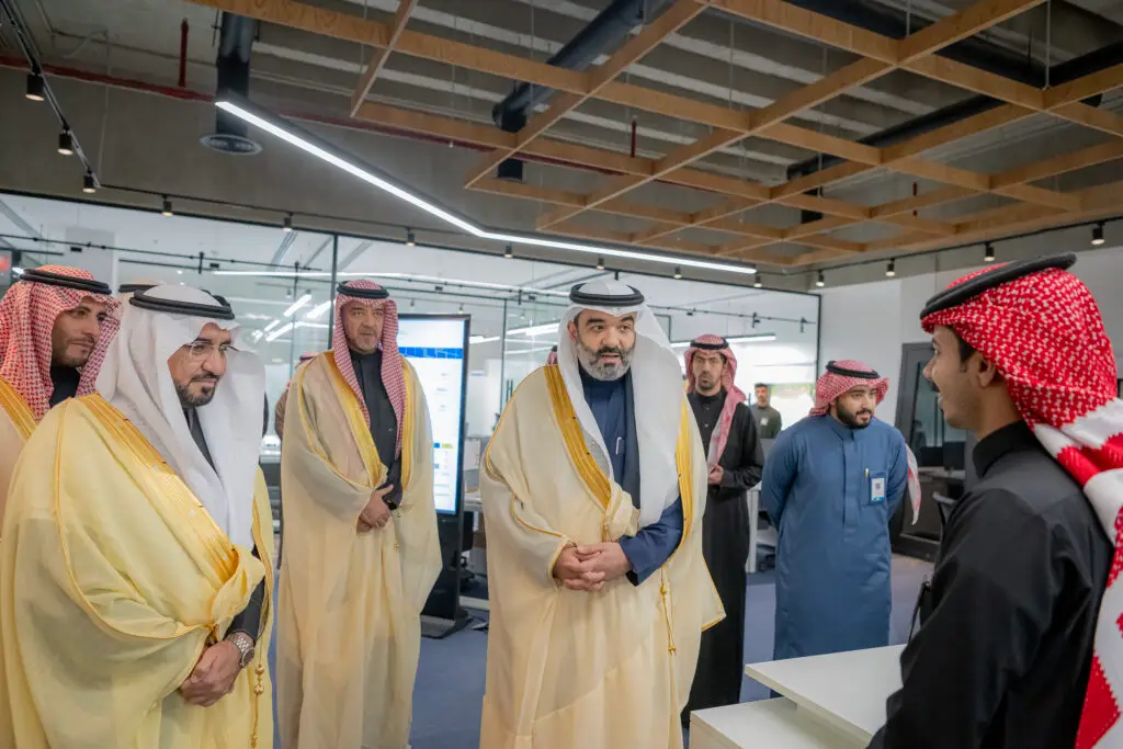 بالتأكيد، إليكم نسخة رسمية ومعاد صياغتها بشكل مناسب بناءً على إرشاداتكم: "مجموعة من الرجال يرتدون الزي السعودي التقليدي، بمن فيهم ممثل من جامعة القصيم يرتدي شماغًا سعوديًا أو غترةً، ينخرطون في نقاش داخل بيئة مكتبية حديثة. يقف شخص واحد مقابل المجموعة على المكتب، ويستمعون باهتمام. يعكس المكان، الذي يتميز بجدران زجاجية وشاشات رقمية، جوًا مخصصًا للبحث والابتكار.